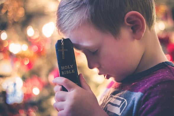 Child holding a Bible with a lit Christmas tree in the background