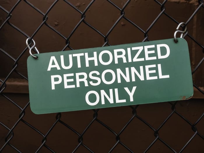 Light green outdoor sign, slightly crooked, on black chain-link fence by dark red steel wall AUTHORIZED PERSONNEL ONLY