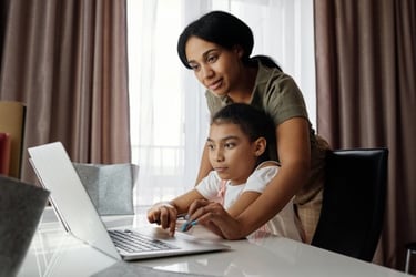 A child is learning on a laptop on a desk. An adult looks over the child's shoulder.